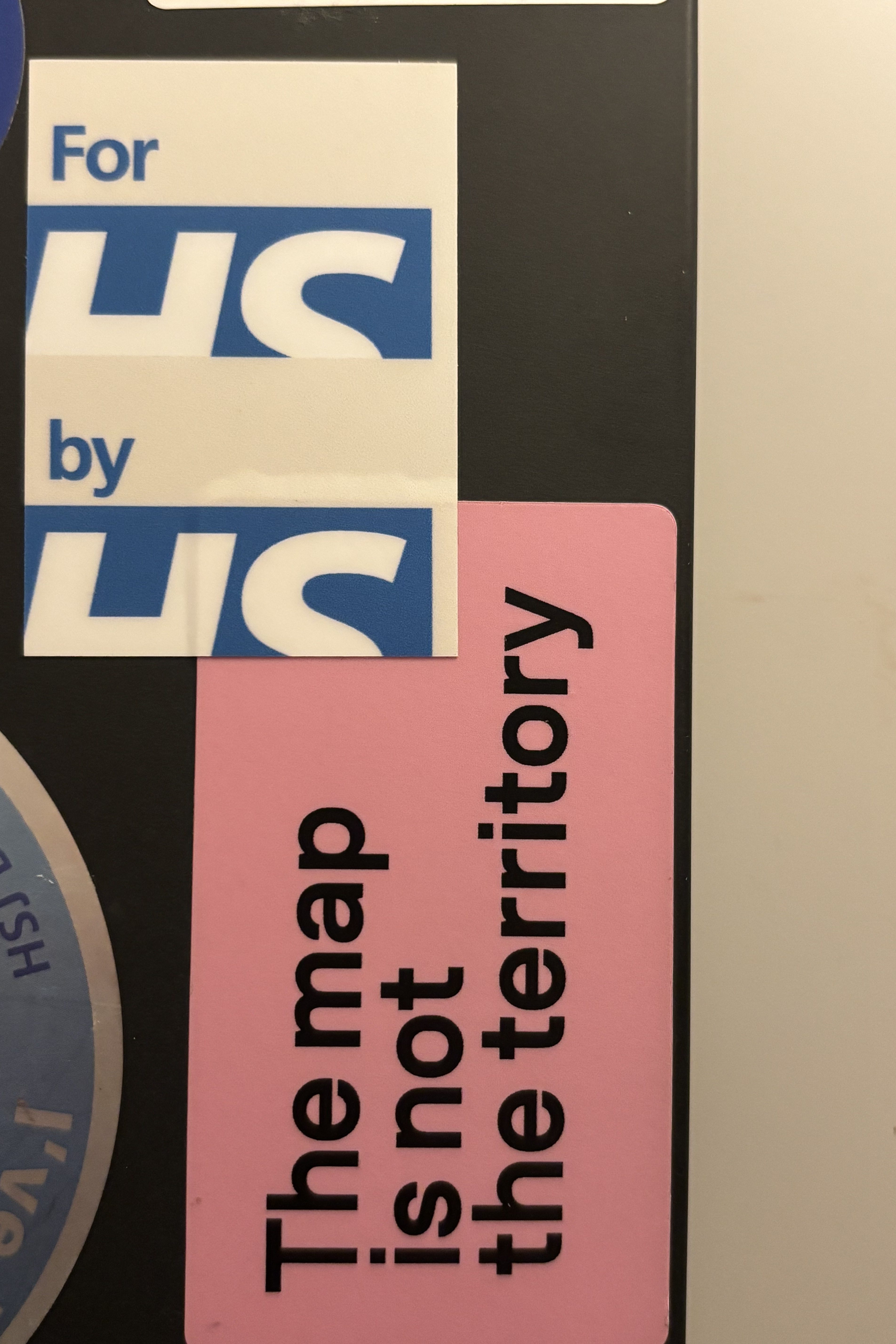 Close-up of two stickers on a black surface. One sticker reads 'For us by us' using the NHS logo and the other reads 'The map is not the territory' on a pink background.